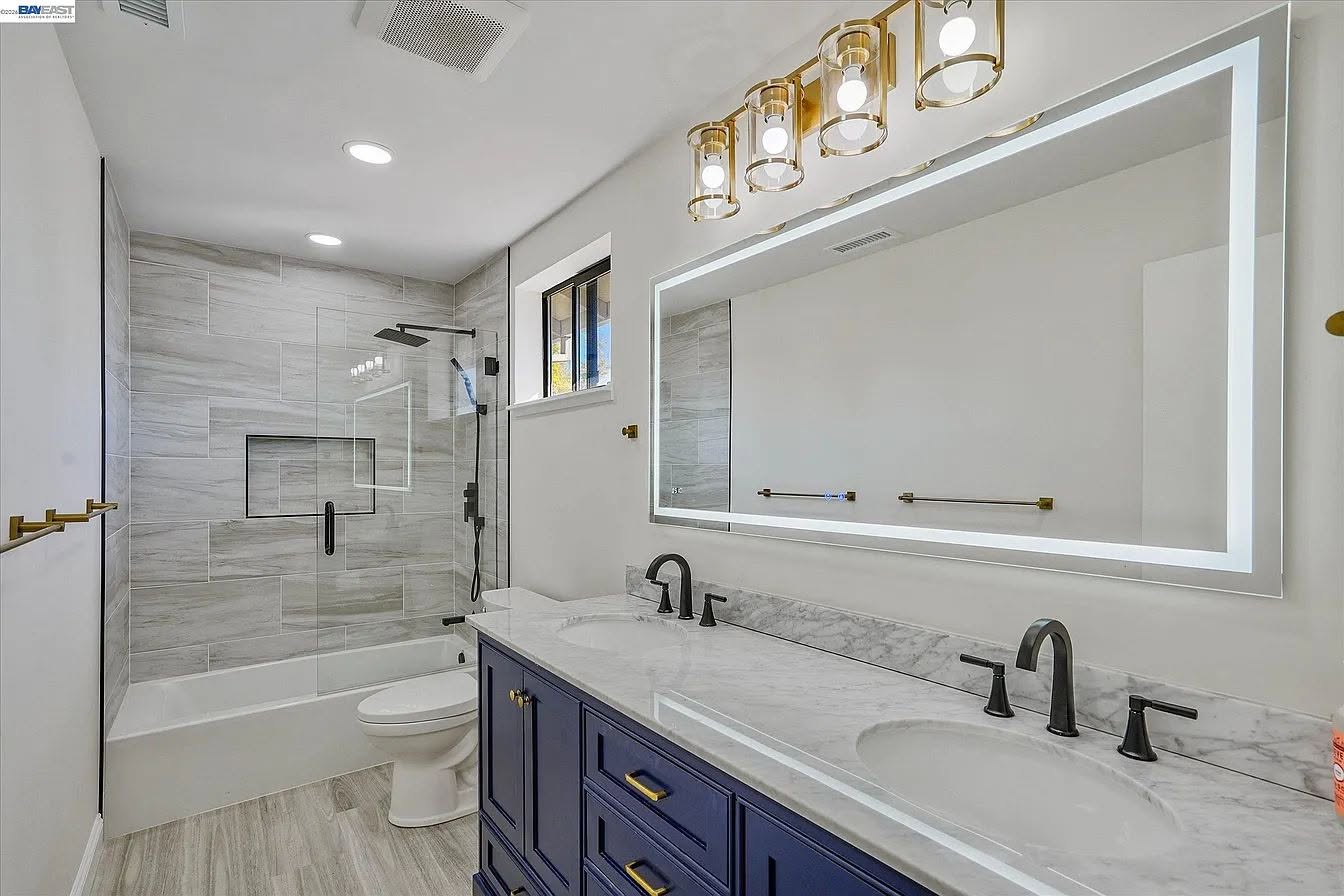 Finished bathroom with navy double vanity, illuminated mirror, and glass tub enclosure